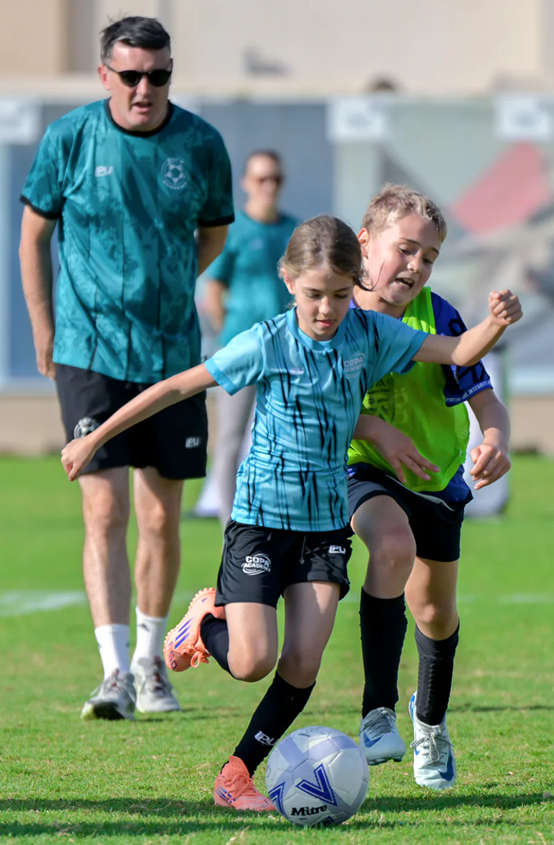 Children compete for the ball during a football academy training session while a coach watches nearby.