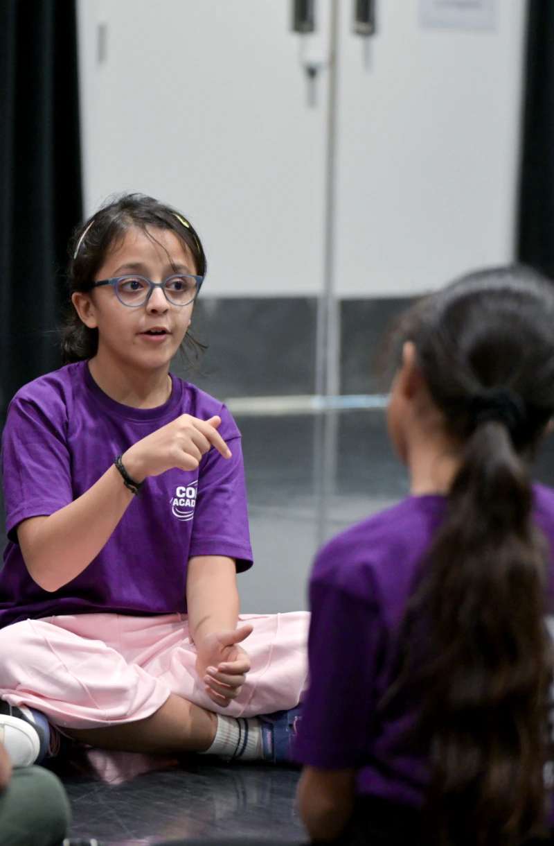 Two children in purple Copa Academy shirts sit on the floor during a class discussion.