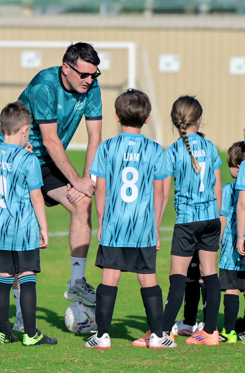 Youth football coach speaking to children in blue kits during a training huddle on the pitch.